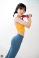 A woman in a yellow top and blue leggings holding a pink dumbbell.